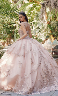 A woman in the Juliet JT1450J Quinceanera Dress, featuring a pale pink ball gown and sweetheart neckline, stands outdoors among lush greenery, glancing over her shoulder.