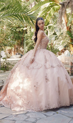 A young woman stands outdoors in a lush garden, wearing the Quinceanera Dress By Juliet JT1450J, featuring a sweetheart neckline, intricate beading, and floral embroidery on voluminous blush pink embroidered tulle.