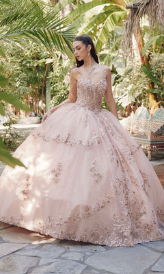 A woman stands outdoors in the Juliet JT1450J Quinceanera Dress, a light pink ball gown with a sweetheart neckline and intricate floral details, surrounded by lush green plants and trees.