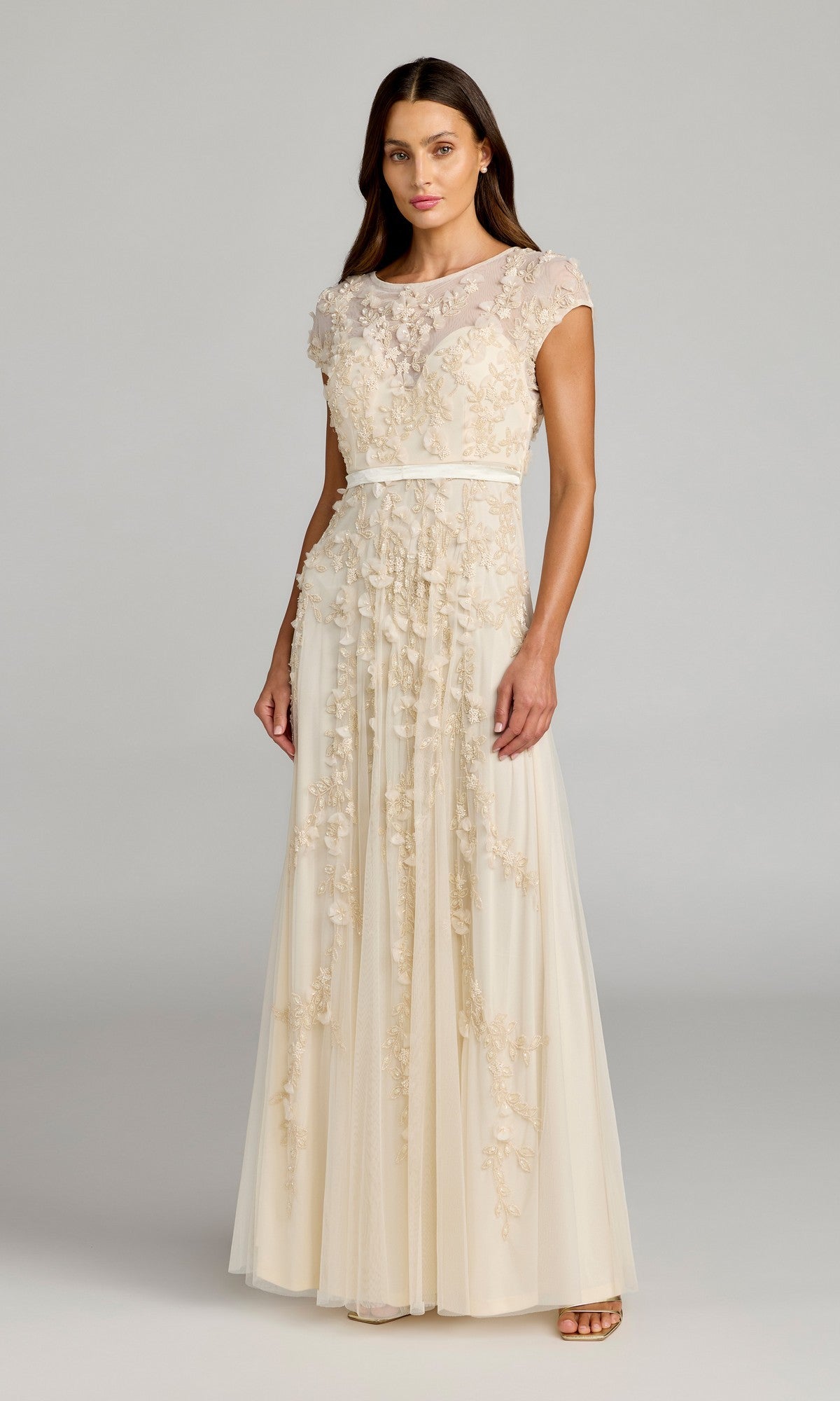 A woman wears the Mac Duggal 14521 Cap-Sleeve Long Formal Dress in cream with floral embroidery and a ribbon belt, making it ideal for special occasions, set against a simple gray background.