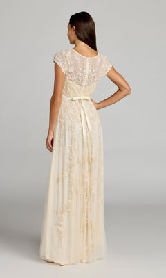A woman stands facing away, wearing the Mac Duggal 14521 Cap-Sleeve Long Formal Dress in cream, featuring floral lace accents and a ribbon waist tie.