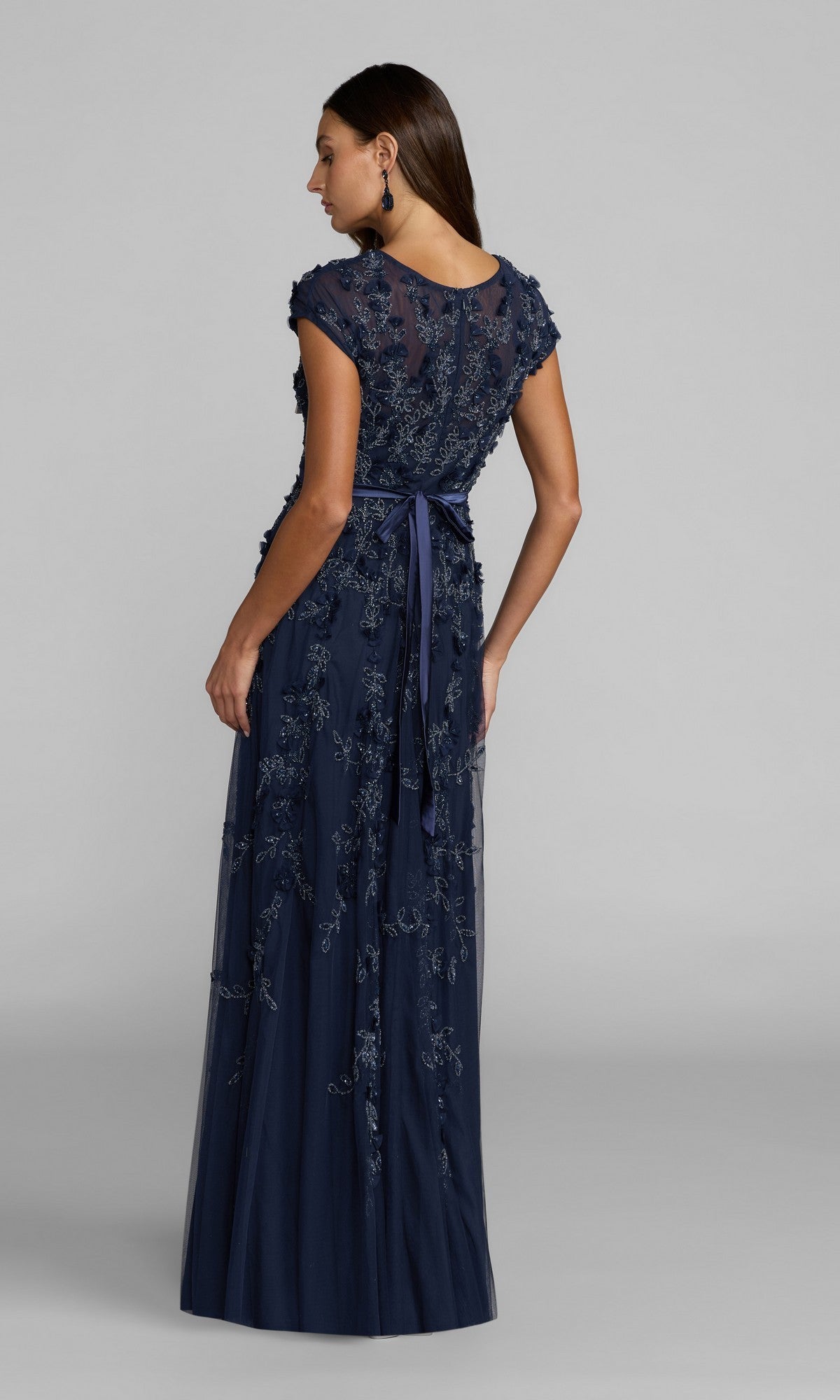 A woman faces away, wearing the Mac Duggal 14521—a floor-length, navy blue cap-sleeve formal dress featuring floral embroidery and a waist ribbon.
