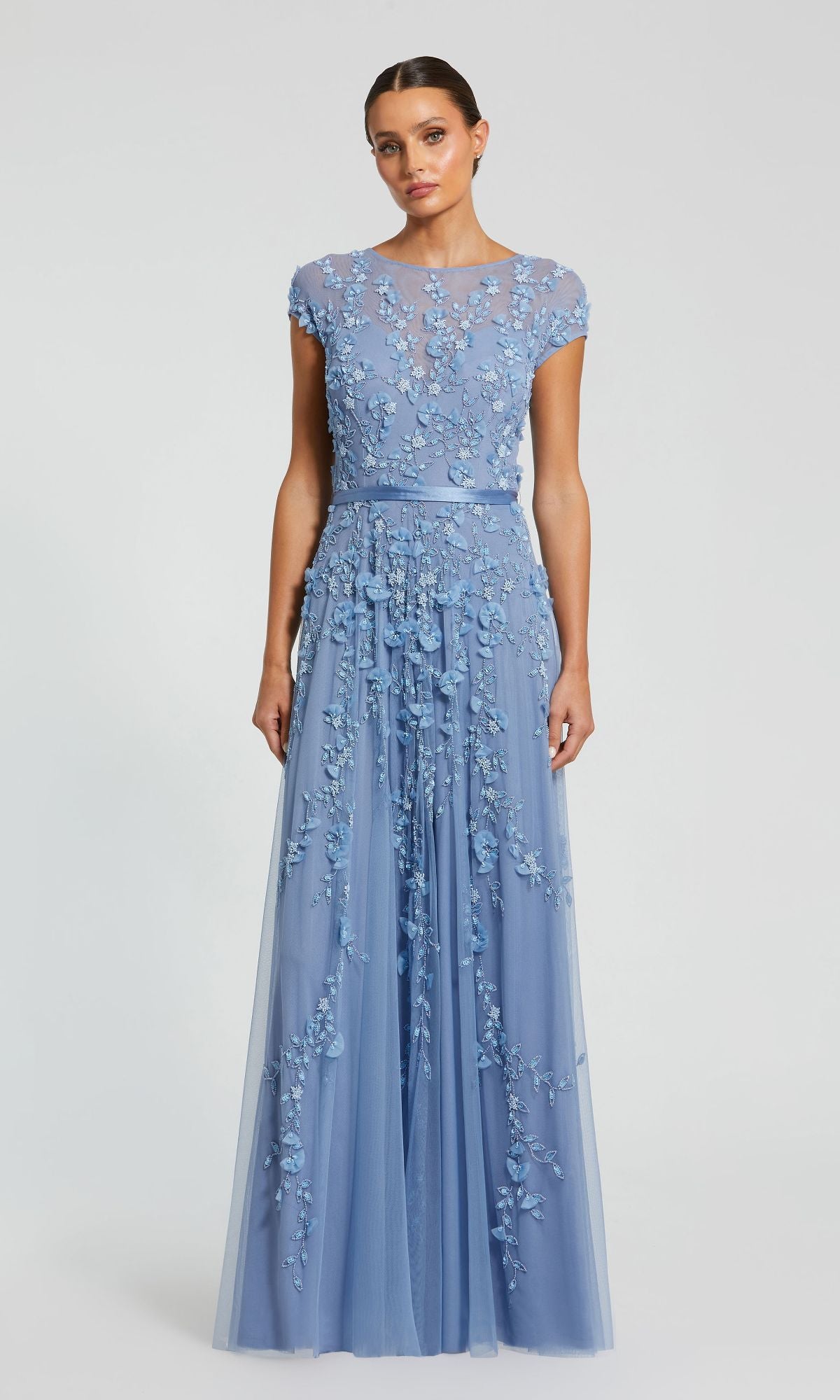 A woman wears the Long Sky Blue Formal Dress: Mac Duggal 14521, a floor-length gown with cap sleeves, floral embroidery, and a sheer overlay, standing against a plain background.