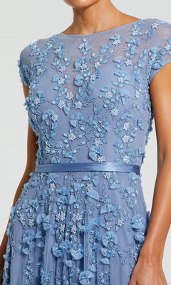 A woman wearing the Mac Duggal 14521— a long sky blue formal dress featuring short sleeves, 3D floral appliqués, and beaded embellishments.