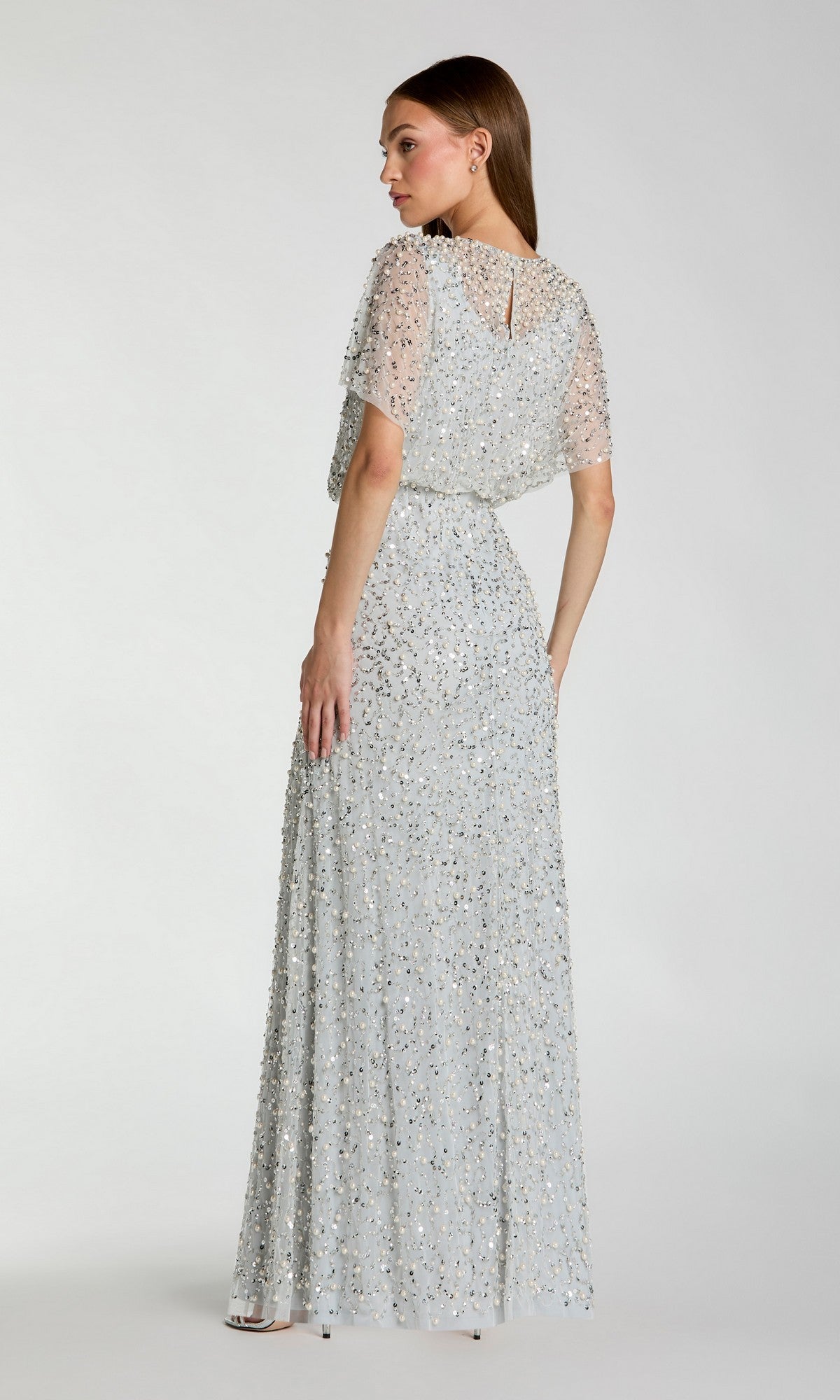 A woman in the Mac Duggal 14523 Beaded Long Nude Evening Gown, featuring short sleeves and floor-length design, stands facing slightly away against a plain background.