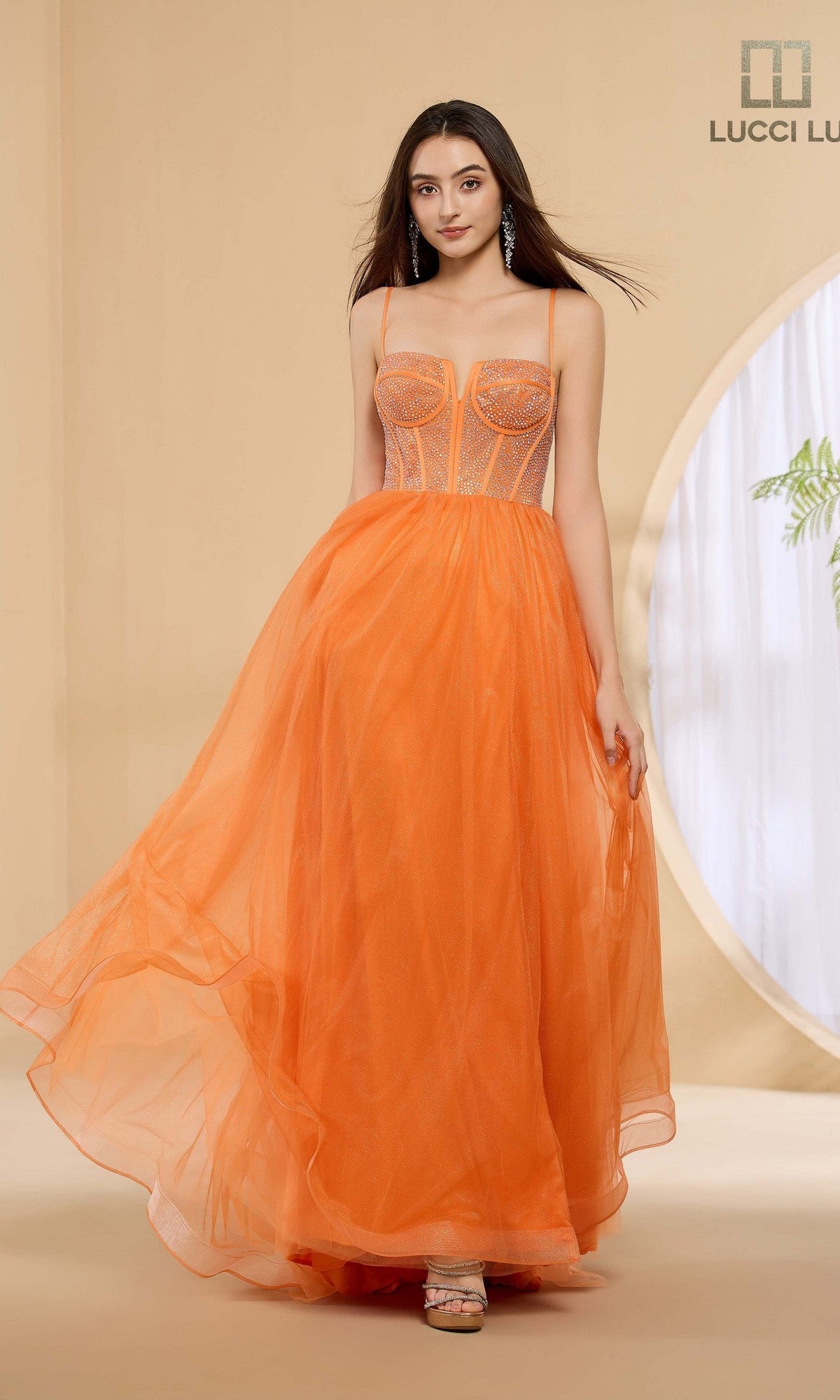 A woman in the Lucci Lu 1457 long orange prom dress with a rhinestone corset bodice and spaghetti straps stands indoors against a beige background.