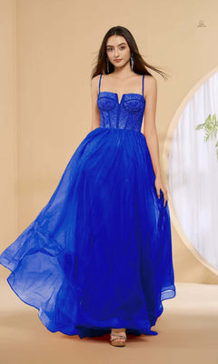 A woman stands indoors on a beige floor, looking at the camera, wearing the Lucci Lu 1457 long prom dress in royal blue with a sleeveless rhinestone corset bodice and tulle A-line skirt.