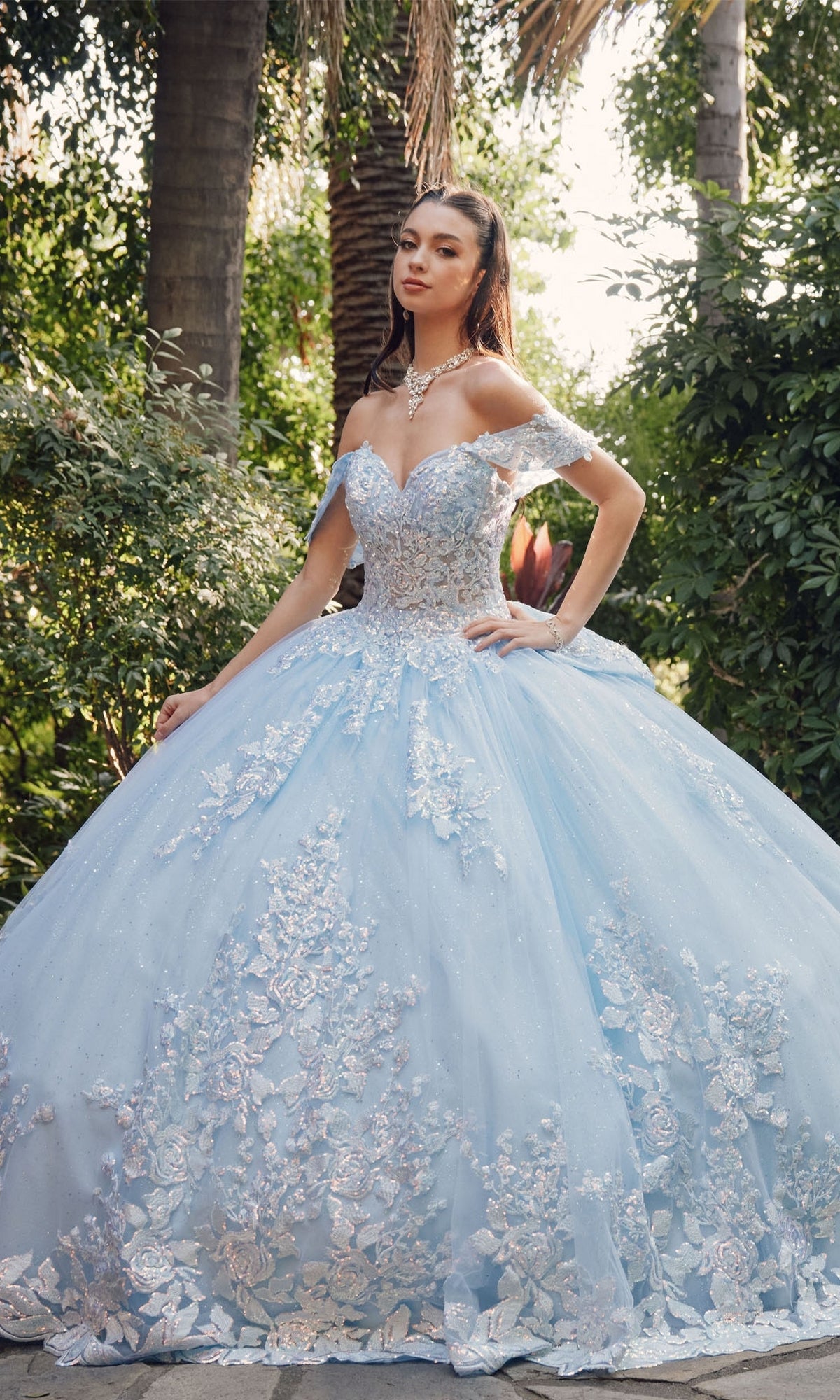 A woman stands in a lush garden wearing the Quinceanera Dress By Juliet JT1458J—an ornate, off-the-shoulder ball gown with floral embroidery and a voluminous light blue skirt.
