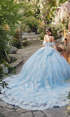 A woman wearing the Quinceanera Dress By Juliet JT1458J, an off-the-shoulder ball gown with a long lace train, stands on a stone path surrounded by lush tropical plants.
