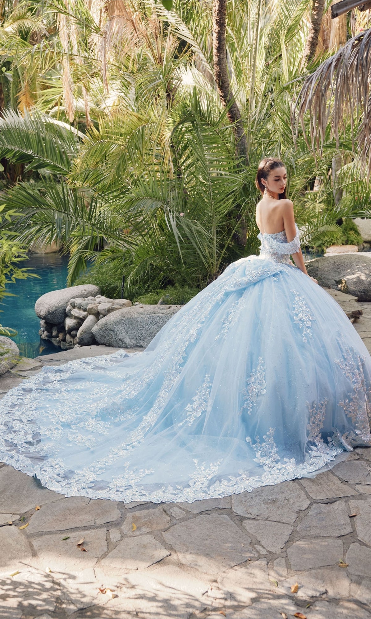 A woman wearing the Quinceanera Dress By Juliet JT1458J, a light blue off-the-shoulder ball gown with a long lace train, stands on a stone path in a lush garden near a pool.