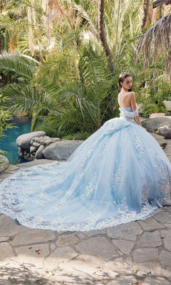 A woman wearing the Quinceanera Dress By Juliet JT1458J, a light blue off-the-shoulder ball gown with a long lace train, stands on a stone path in a lush garden near a pool.