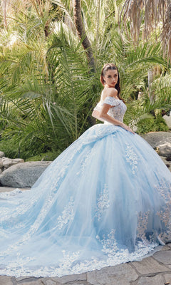A woman stands outdoors before tropical plants and rocks, wearing the Quinceanera Dress By Juliet JT1458J—a blue, off-the-shoulder ball gown with floral lace details.