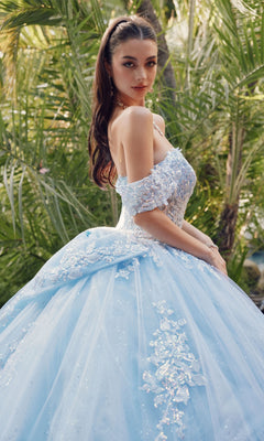 A woman stands outdoors before green foliage, wearing the Quinceanera Dress By Juliet JT1458J—a light blue, off-the-shoulder ball gown adorned with lace and floral details.
