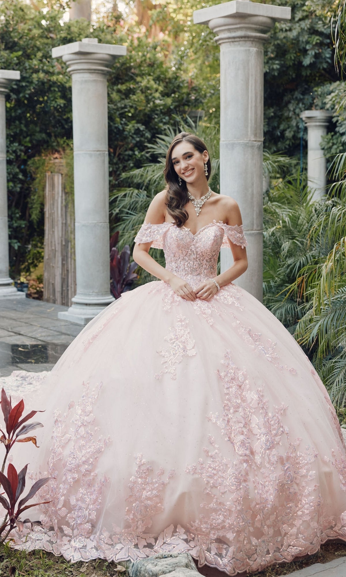 A woman in the Juliet JT1458J Quinceanera Dress, a pink off-the-shoulder ball gown with floral details, stands in a garden with stone columns and lush greenery.