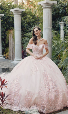 A woman in the Juliet JT1458J Quinceanera Dress, a pink off-the-shoulder ball gown with floral details, stands in a garden with stone columns and lush greenery.