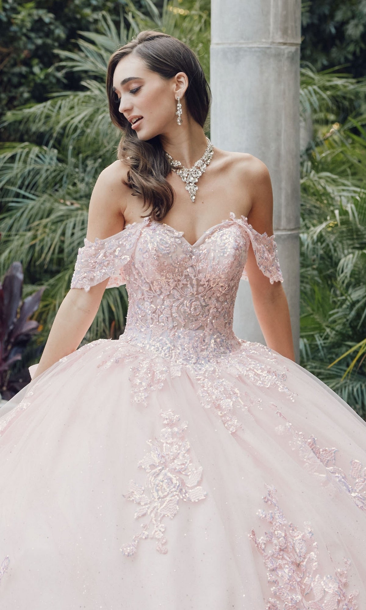 A woman wearing the Quinceanera Dress By Juliet JT1458J, a sparkly off-the-shoulder ball gown with floral details, stands outdoors accessorized with a necklace and earrings.