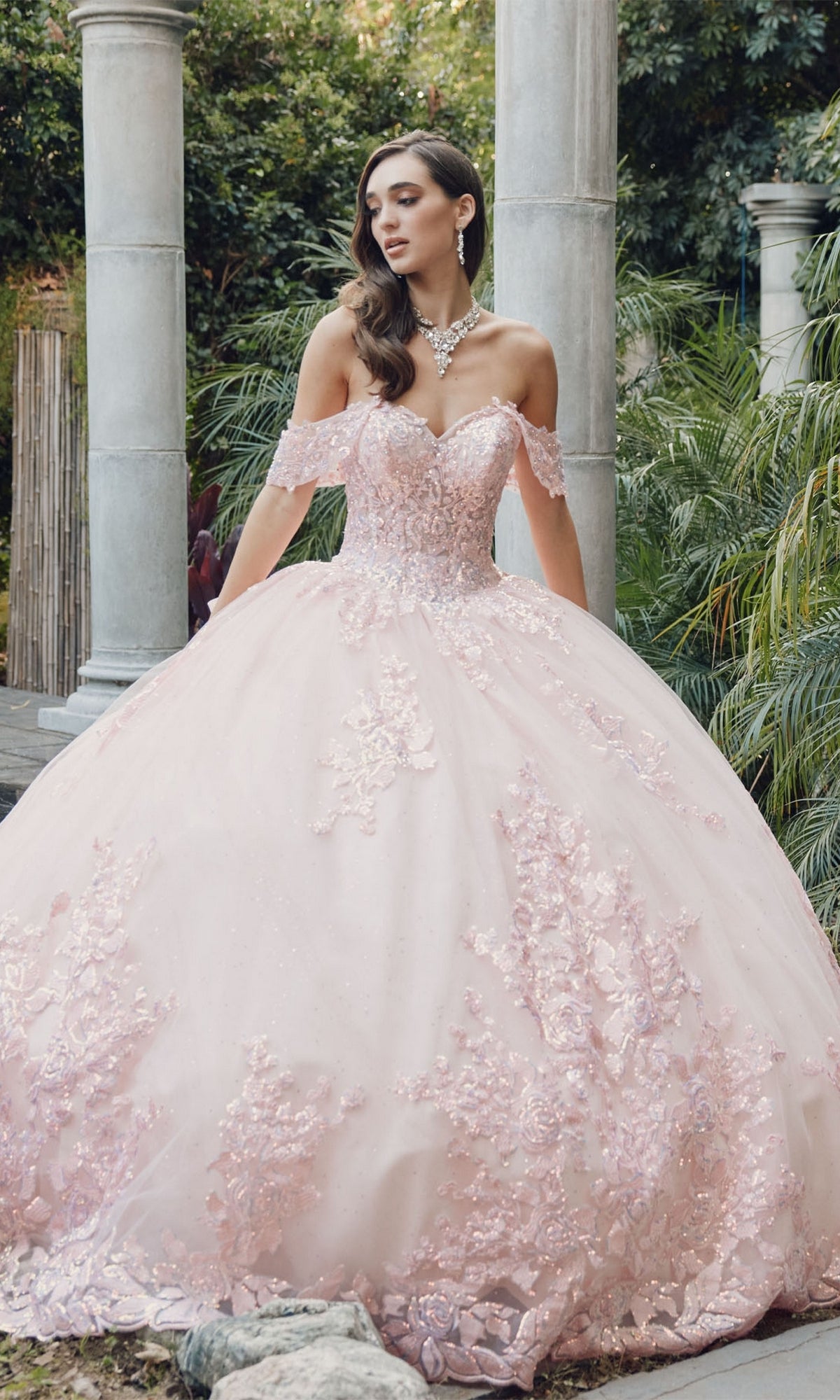 A woman stands outdoors in the Quinceanera Dress By Juliet JT1458J, an off-the-shoulder light pink tulle A-line gown with floral details, jewelry, and surrounded by greenery and stone columns.
