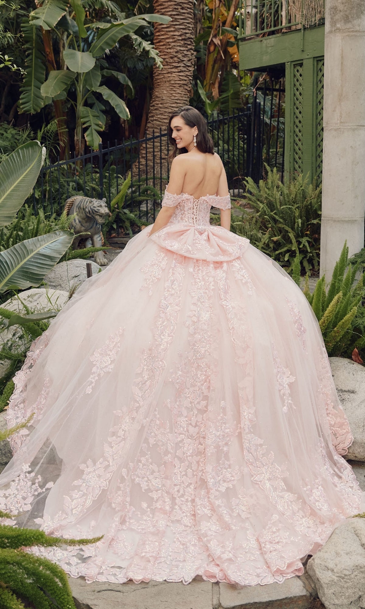 A woman stands outdoors among lush green plants, wearing the Quinceanera Dress By Juliet JT1458J—a pale pink, off-shoulder ball gown featuring floral lace details and a full, layered skirt.