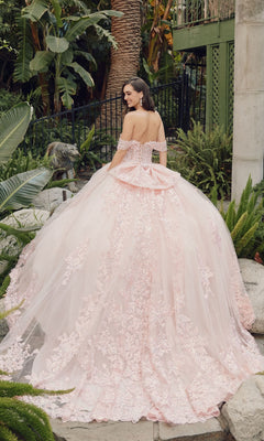 A woman stands outdoors among lush green plants, wearing the Quinceanera Dress By Juliet JT1458J—a pale pink, off-shoulder ball gown featuring floral lace details and a full, layered skirt.