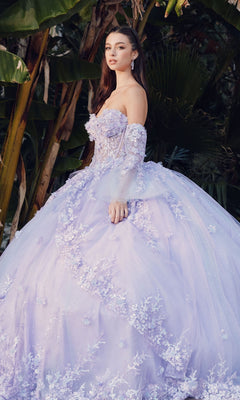 A woman wearing the Quinceanera Dress JT1459J by Juliet, a lilac strapless tulle A-line gown with floral appliqués, stands outdoors in front of lush green leaves as the elegant dress flows around her.