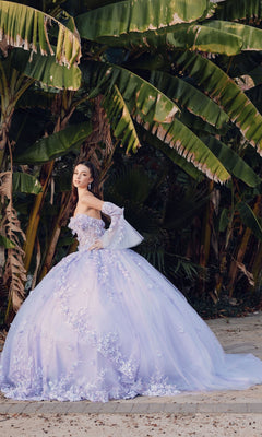 A woman in the Quinceanera Dress JT1459J by Juliet, an off-the-shoulder gown with floral details, stands on a path in front of large green banana leaves.
