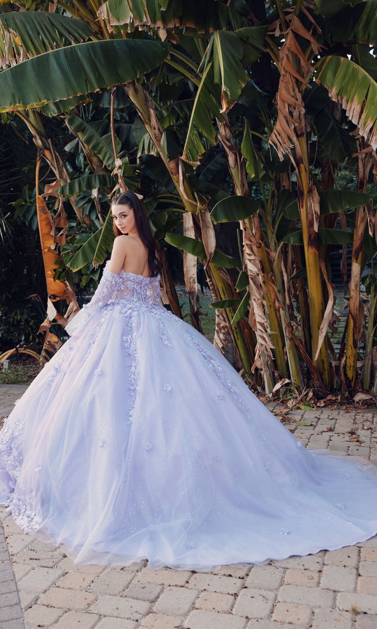 A woman in the Quinceanera Dress JT1459J by Juliet stands on a stone path, turning to look back, with large green banana leaves in the background.