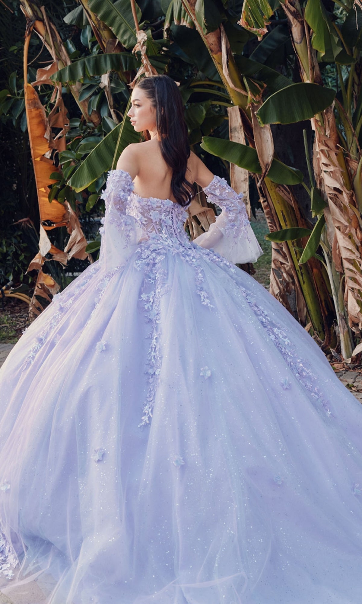 A woman stands outdoors before leafy plants, wearing the Quinceanera Dress JT1459J by Juliet, featuring off-the-shoulder sleeves, floral appliqués, and a full tulle skirt in lilac.