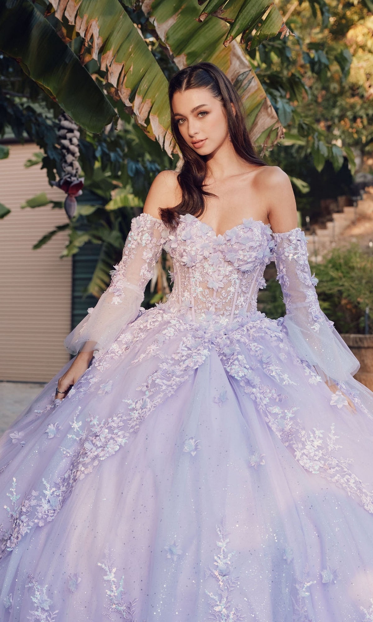 A woman stands outdoors in the Quinceanera Dress JT1459J by Juliet—an off-the-shoulder lilac ball gown with floral embellishments and sheer sleeves—elegantly framed by lush greenery.
