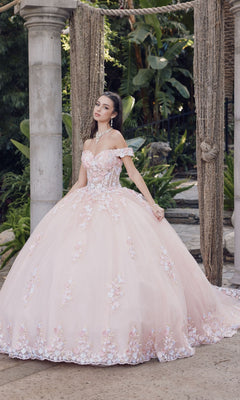 A woman stands outdoors in the Quinceanera Dress By Juliet JT1460J, a blush pink, off-the-shoulder ball gown with floral embellishments and a full skirt, surrounded by greenery and stone pillars.