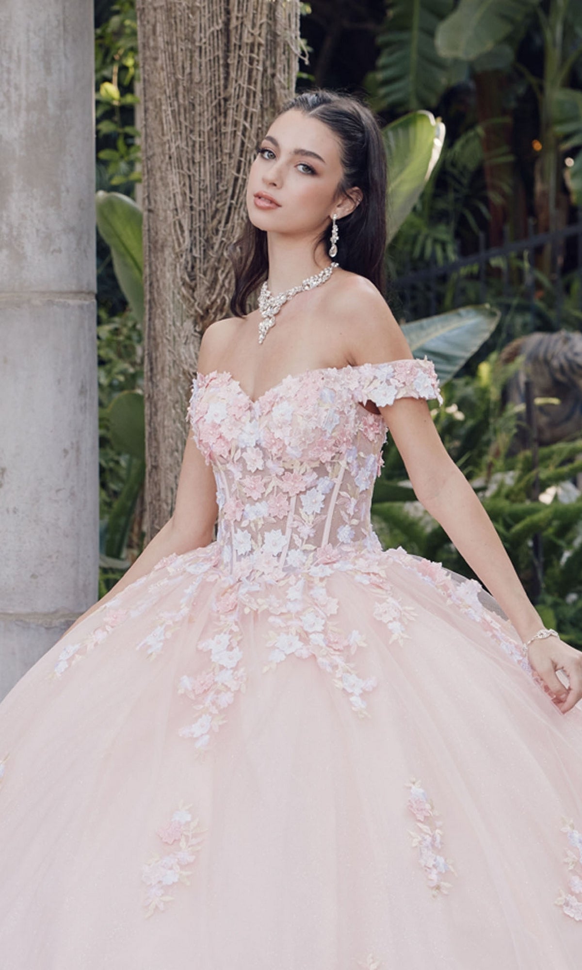A woman stands outdoors near trees and plants wearing the Quinceanera Dress By Juliet JT1460J, a blush pink off-the-shoulder ball gown featuring floral embellishments.