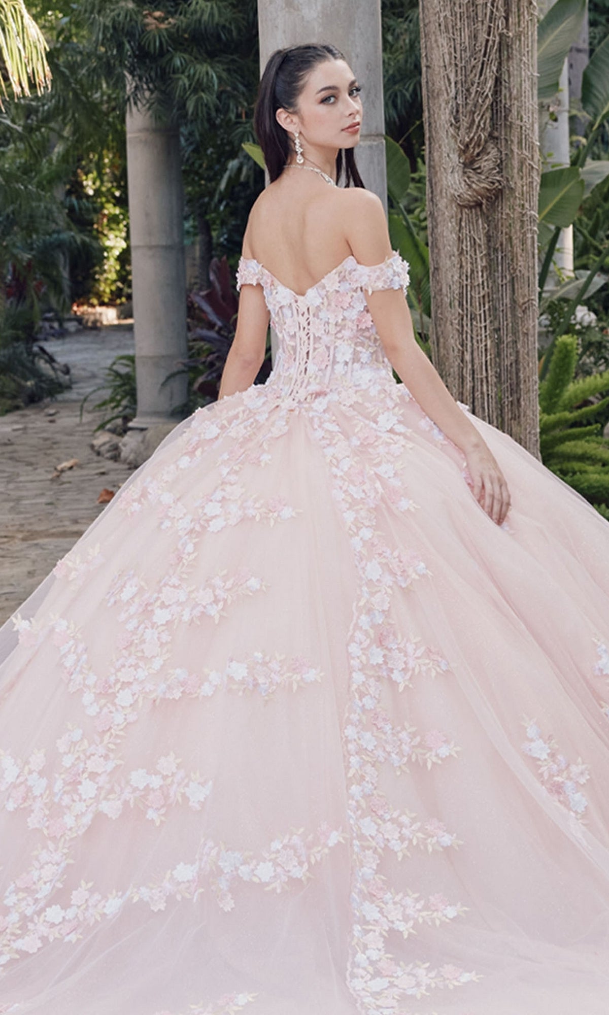 Wearing the Quinceanera Dress By Juliet JT1460J in blush pink with floral appliqués, a woman stands outdoors amid lush greenery, glancing over her shoulder in an elegant off-the-shoulder ball gown.