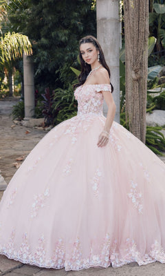 A woman stands outdoors in the Quinceanera Dress By Juliet JT1460J—a blush pink, off-the-shoulder ball gown with floral embellishments and a full skirt—surrounded by greenery and stone columns.