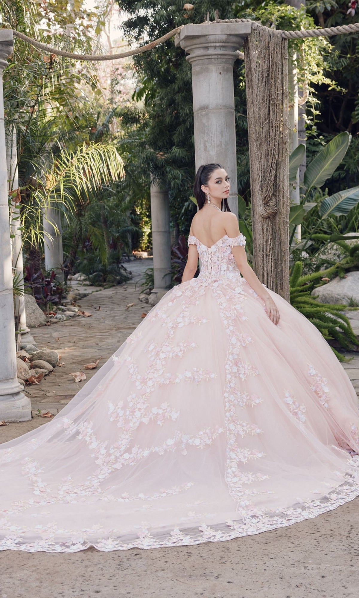 A woman wearing the Juliet JT1460J blush pink off-the-shoulder quinceanera dress with floral appliqués stands in a garden surrounded by stone columns and lush greenery.