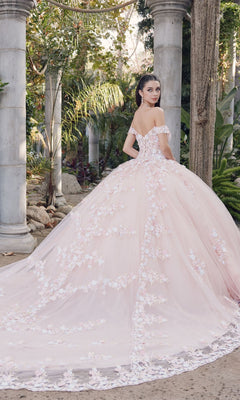 A woman wearing the Quinceanera Dress By Juliet JT1460J in blush pink, featuring off-the-shoulder floral appliqués, stands outdoors near stone columns and greenery, looking over her shoulder.