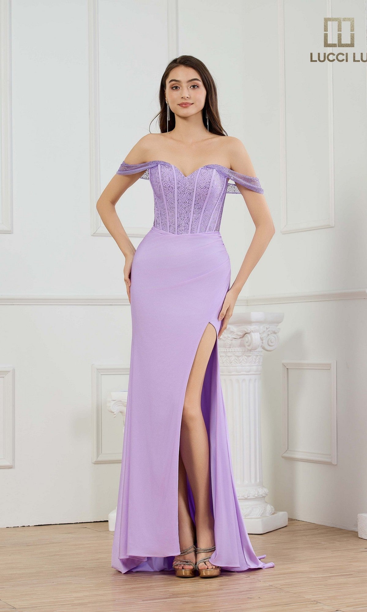A woman poses indoors with hands on her hips in front of a white pillar, wearing the Lucci Lu 1466 long lilac prom dress featuring an off-the-shoulder design, fitted corset bodice, and high slit.