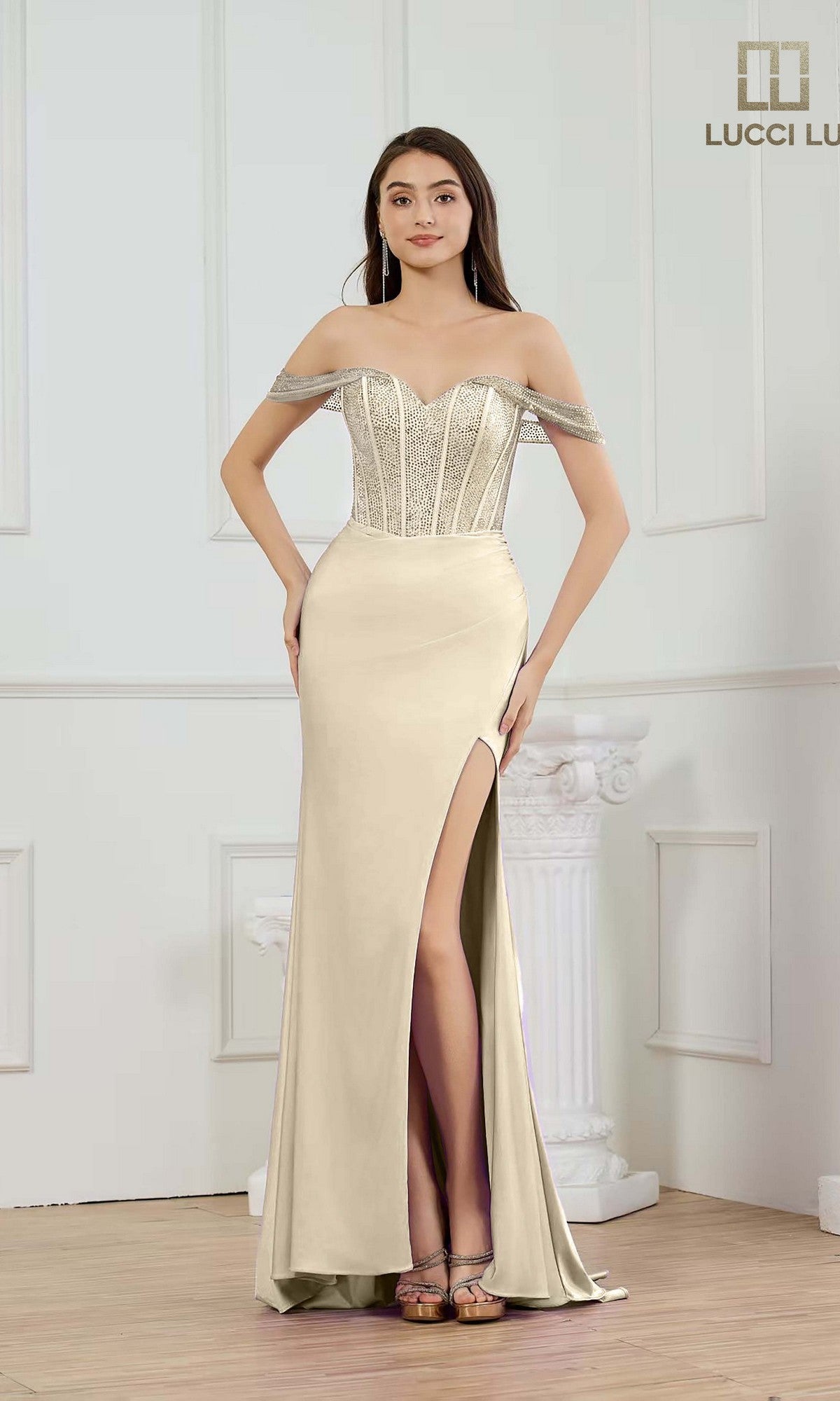 A woman poses indoors against white paneled walls and a column wearing the gold Lucci Lu 1466 long prom dress, featuring an off-the-shoulder neckline, fitted corset bodice, glitter sheath skirt, and silver embellishments.