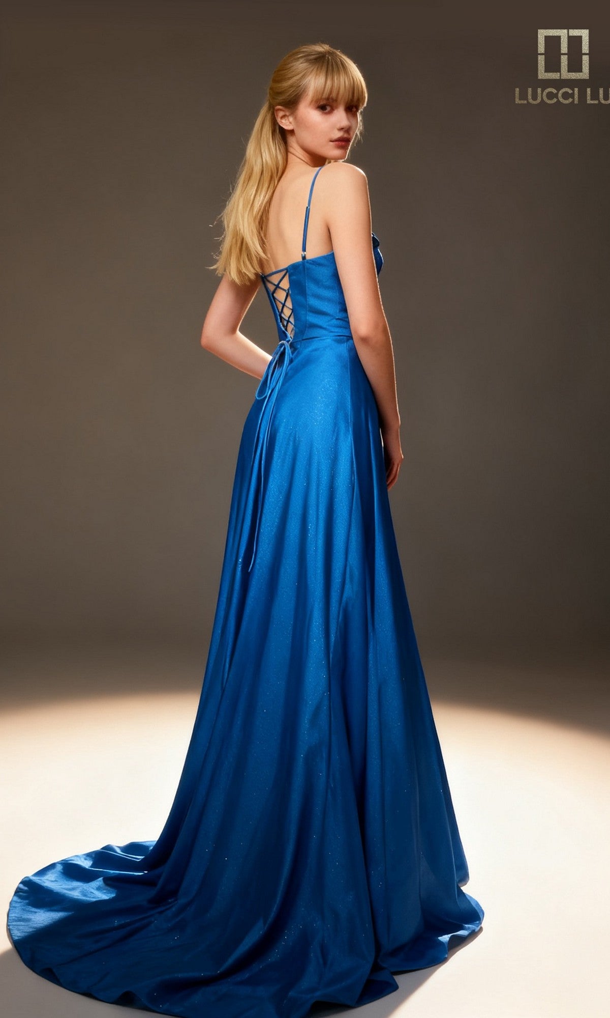 A woman with long blonde hair stands facing away in a studio, wearing the Lucci Lu 1477 Long Prom Dress in blue, featuring an elegant satin a-line silhouette with a lace-up back.