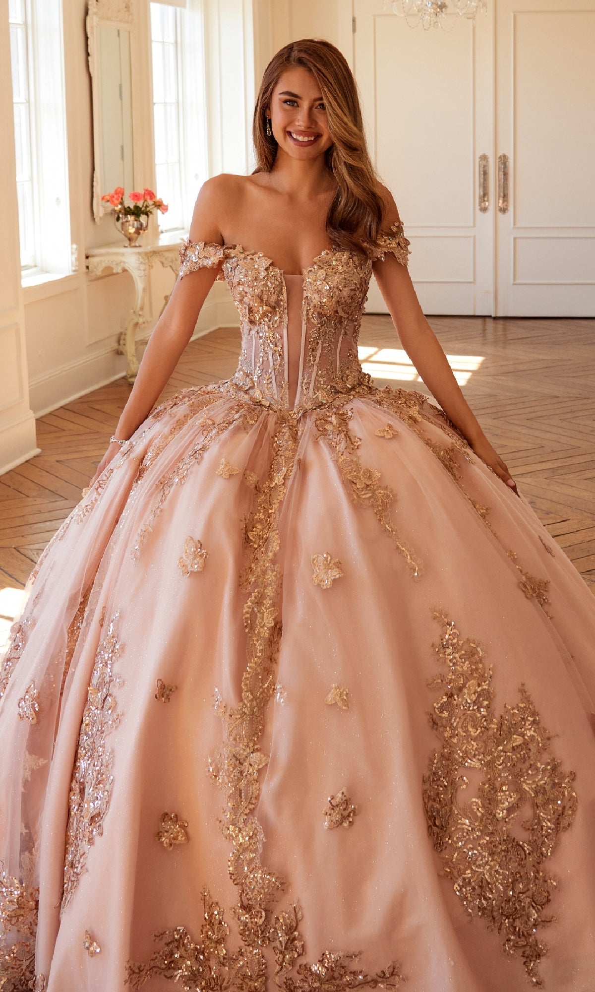 A woman wears the Quinceanera Dress JT1485J By Juliet, a light pink off-the-shoulder gown with gold floral embellishments, standing in a bright room with wood floors and white walls.