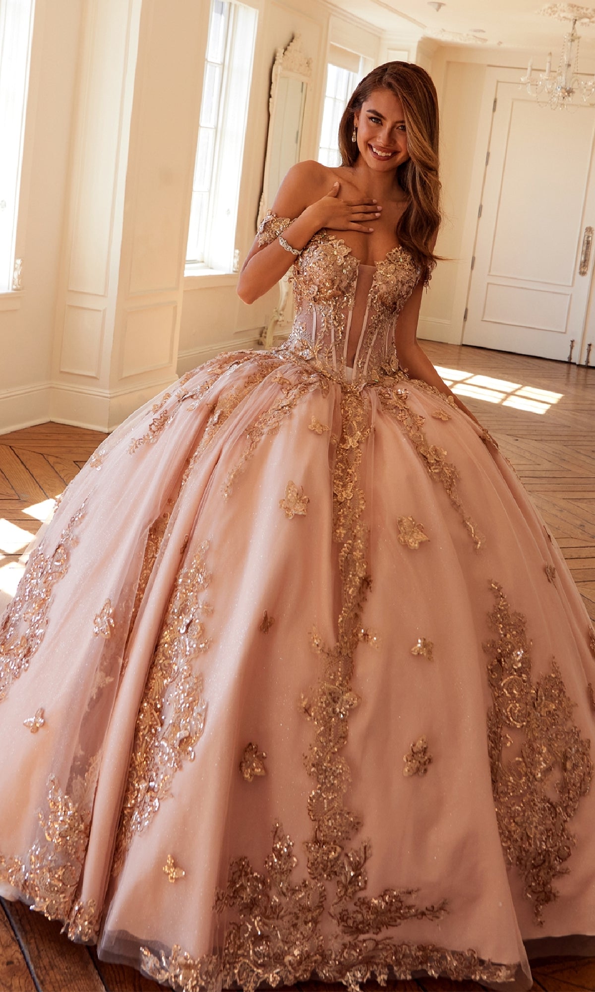 A young woman stands indoors, smiling with a hand to her chest, wearing the Quinceanera Dress JT1485J By Juliet—an off-the-shoulder blush pink ball gown adorned with gold embroidery and floral appliqués.