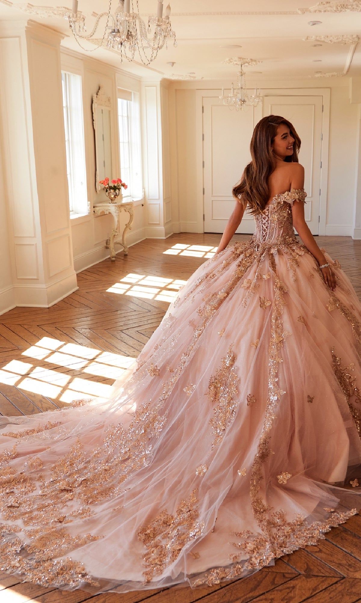 A woman in an elegant room wears the Quinceanera Dress JT1485J By Juliet—an off-the-shoulder blush pink ball gown with gold floral embellishments and a long, flowing train.