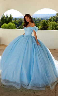 A young woman stands outdoors in the Quinceanera Dress JT1487H By Juliet, featuring a light blue princess style with puff off-the-shoulder sleeves and a full tulle skirt, set against lush greenery and an arched white wall.