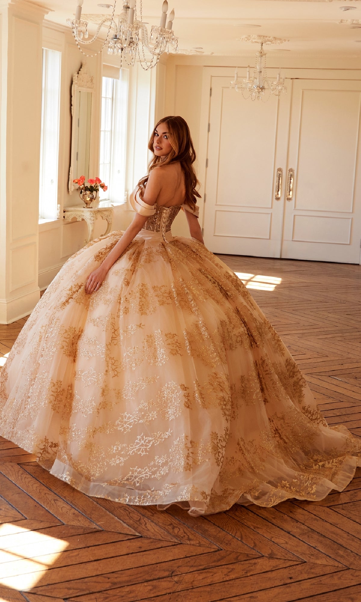 A woman wearing the Quinceanera Dress JT1488H by Juliet—ornate, gold-embroidered, with a corset bodice and ball gown skirt—stands in a bright, elegant room with large windows, wooden floors, and white walls.