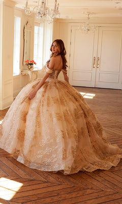 A woman wearing the Quinceanera Dress JT1488H by Juliet—ornate, gold-embroidered, with a corset bodice and ball gown skirt—stands in a bright, elegant room with large windows, wooden floors, and white walls.
