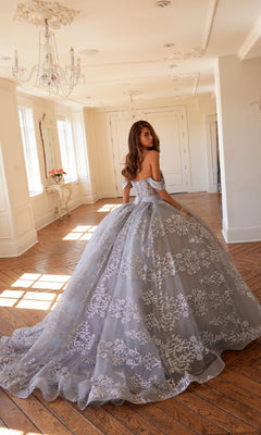 A young woman wearing the Quinceanera Dress JT1488H By Juliet, featuring an off-shoulder corset bodice and floral lace detailing, stands in a bright, elegant room with wooden floors and chandeliers, glancing over her shoulder.