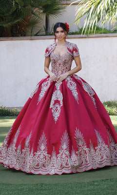 A woman stands outdoors in the Quinceanera Dress 1494 by Dancing Queen, a bright red a-line ball gown with gold lace embroidery, hands clasped at her waist and a matching bolero finishing the look.
