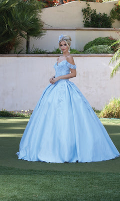 A woman stands on grass wearing the Quinceanera Dress 1501 by Dancing Queen, an off-the-shoulder light blue a-line ball gown with floral details and a matching headpiece. Greenery and a white wall are in the background.