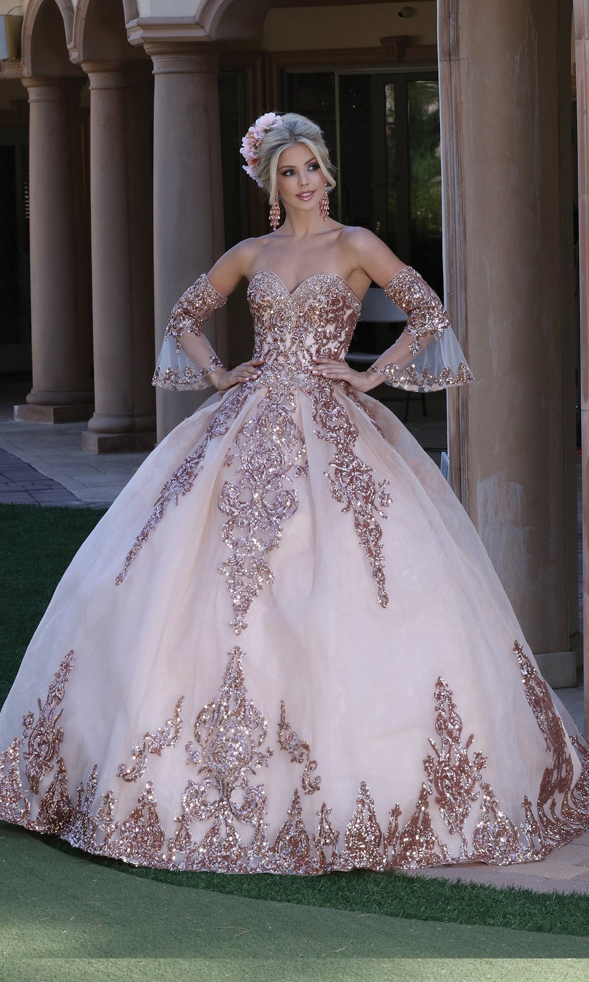 A woman poses outdoors in front of stone columns wearing Dancing Queen’s Quinceanera Dress 1512, an ornate blush pink A-line ball gown with a strapless sweetheart bodice and matching arm accessories.