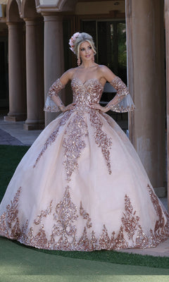 A woman poses outdoors in front of stone columns wearing Dancing Queen’s Quinceanera Dress 1512, an ornate blush pink A-line ball gown with a strapless sweetheart bodice and matching arm accessories.