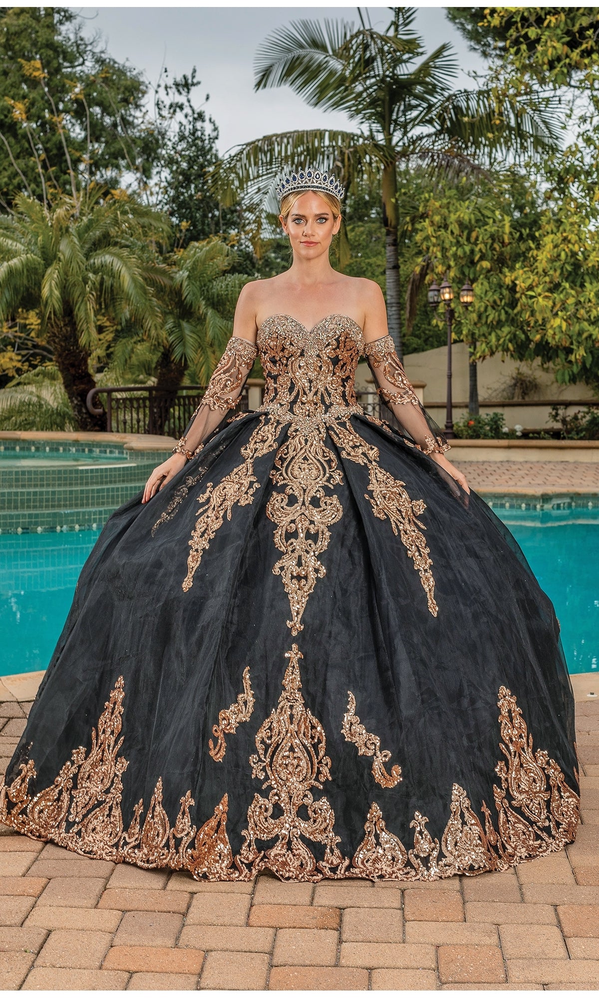 Wearing the Quinceanera Dress 1512 by Dancing Queen—a black and gold a-line ball gown with a strapless sweetheart bodice and off-the-shoulder sleeves—a person stands by a pool in a silver crown, with palm trees and greenery behind.