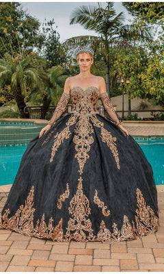 Wearing the Quinceanera Dress 1512 by Dancing Queen—a black and gold a-line ball gown with a strapless sweetheart bodice and off-the-shoulder sleeves—a person stands by a pool in a silver crown, with palm trees and greenery behind.